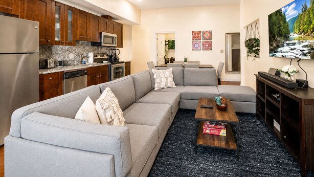 Uma sala de estar moderna com um sofá seccional, uma mesa de centro e uma área de cozinha com armários de madeira e micro-ondas.