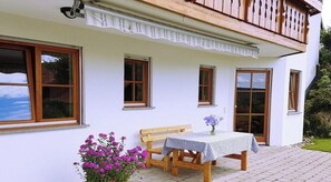 A white building with a balcony, a wooden bench, a table with a checkered tablecloth, and a vase with flowers. - Holiday apartment with 1 bedroom (Missen-Wilhams)