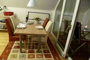 Ein Essbereich mit einem Tisch für zwei Personen, Stühlen, einer Blumenvase und einer Glastür, die auf einen Balkon führt.