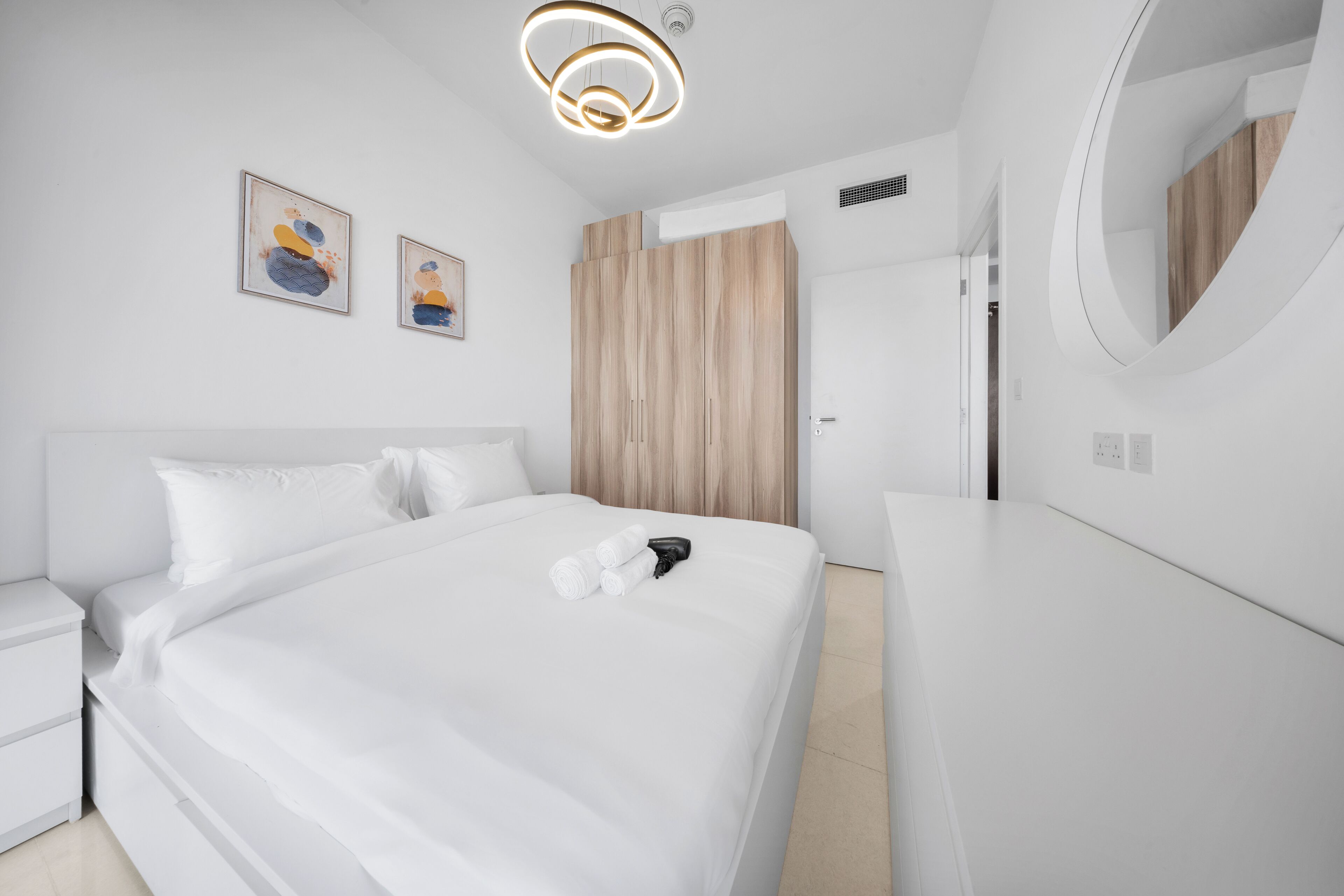A modern hotel room with a large bed, a wooden wardrobe, a round mirror, and two framed artworks on the wall.