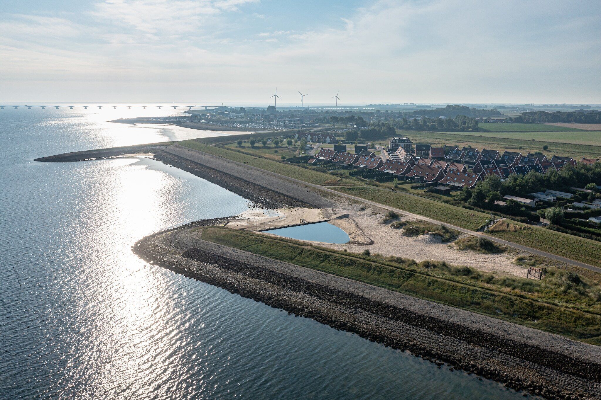 An aerial view of a riverside town with a bridge, wind turbines, and a row of houses.