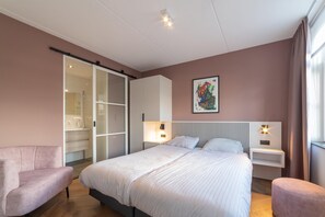 A modern bedroom with a bed, two bedside tables, a nightstand, a sofa, and a sliding glass door leading to a bathroom.