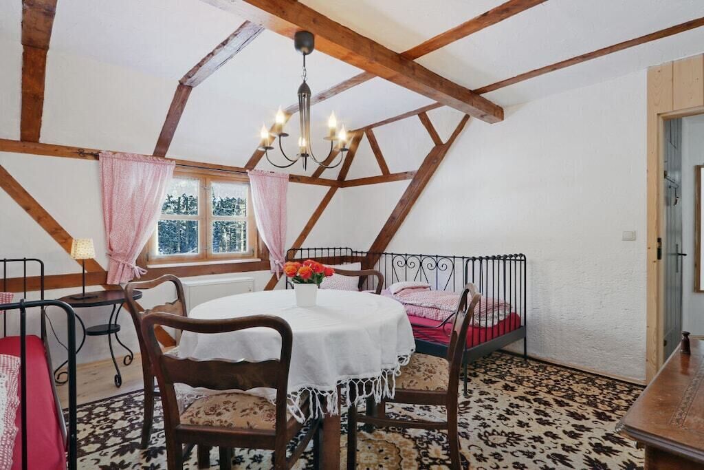 A room with a high ceiling, exposed wooden beams, a round table with chairs, a bed with a metal frame, and a window with curtains.