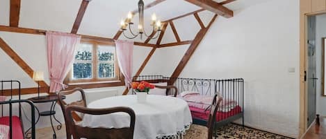 A room with a high ceiling, exposed wooden beams, a round table with chairs, a bed with a metal frame, and a window with curtains.