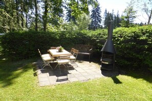 An outdoor patio with a table and chairs, surrounded by lush greenery and a chimney. - Holiday home with 3 bedrooms (Auerbach OT Grünheide)