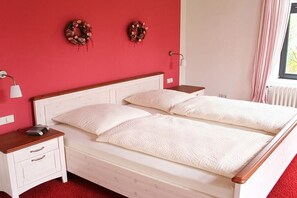 Una cama doble con ropa de cama blanca, una mesita de noche con lámpara y dos coronas decorativas colgadas en la pared.