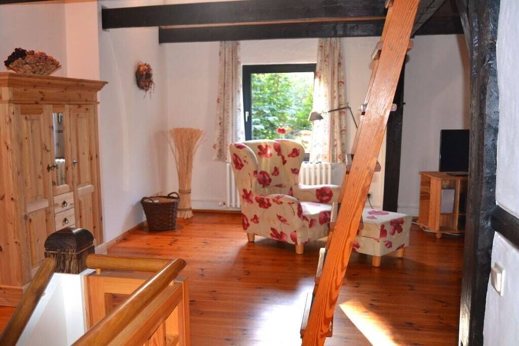 Une pièce avec un plancher en bois, un fauteuil à motifs floraux, un meuble en bois et une fenêtre avec des rideaux.