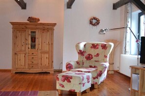 Un mueble de madera con cajones, un sillón floral y un reposapiés a juego en una habitación con suelo de madera y una ventana.