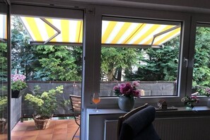 A room with a view of trees through large windows, a chair, a table with a glass, and a small house model. - Holiday apartment with 2 bedrooms (Lübeck)