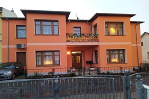Ein zweistöckiges Gebäude mit Balkon, einem geparkten Auto davor und einem Metallzaun.