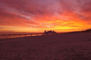 Ein Strand mit einem Steg, der sich ins Wasser erstreckt, ein Gebäude mit weißer Fassade und ein Sonnenuntergangshimmel.