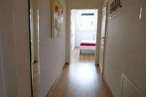Een smalle gang met een bed aan het einde, een schilderij aan de muur en een raam met zonwering.