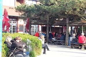 An outdoor seating area with tables and chairs, surrounded by greenery and buildings with red awnings. - Achtern Diek “Sonneneck” (Zingst)