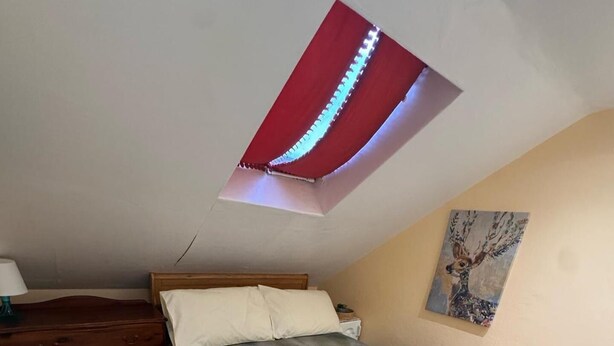 Un dormitorio con cama, cómoda de madera, una mesita, un cuadro en la pared y un tragaluz.