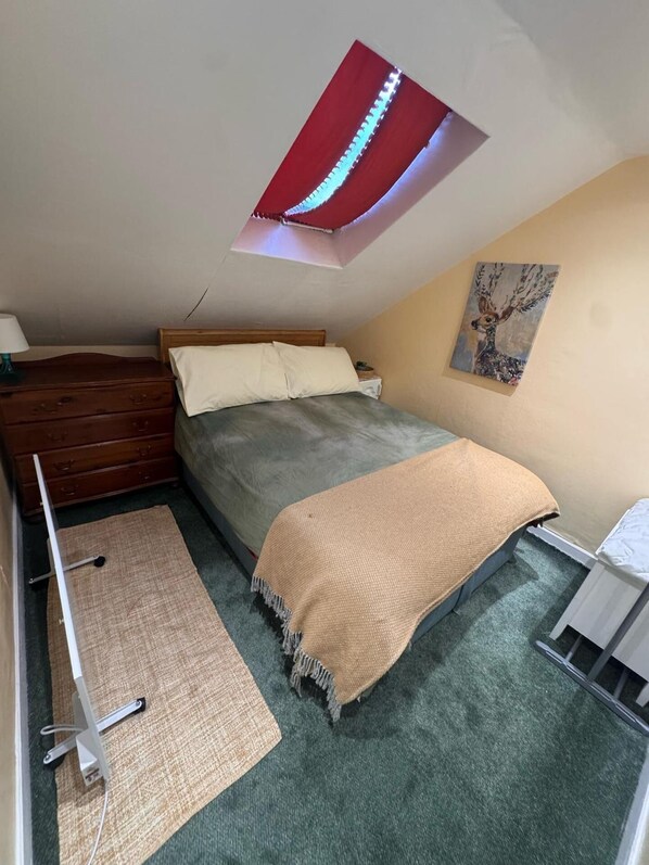 Un dormitorio con cama, cómoda de madera, una mesita, un cuadro en la pared y un tragaluz.