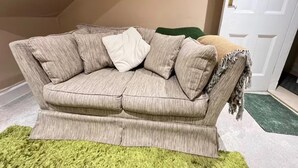 Un sofá beige con una manta y almohadones, sobre una alfombra verde en una habitación con una puerta blanca y un cuadro en la pared.