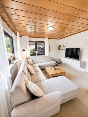 A modern living room with a sectional sofa, a coffee table, a flat-screen TV, and a large window with curtains.