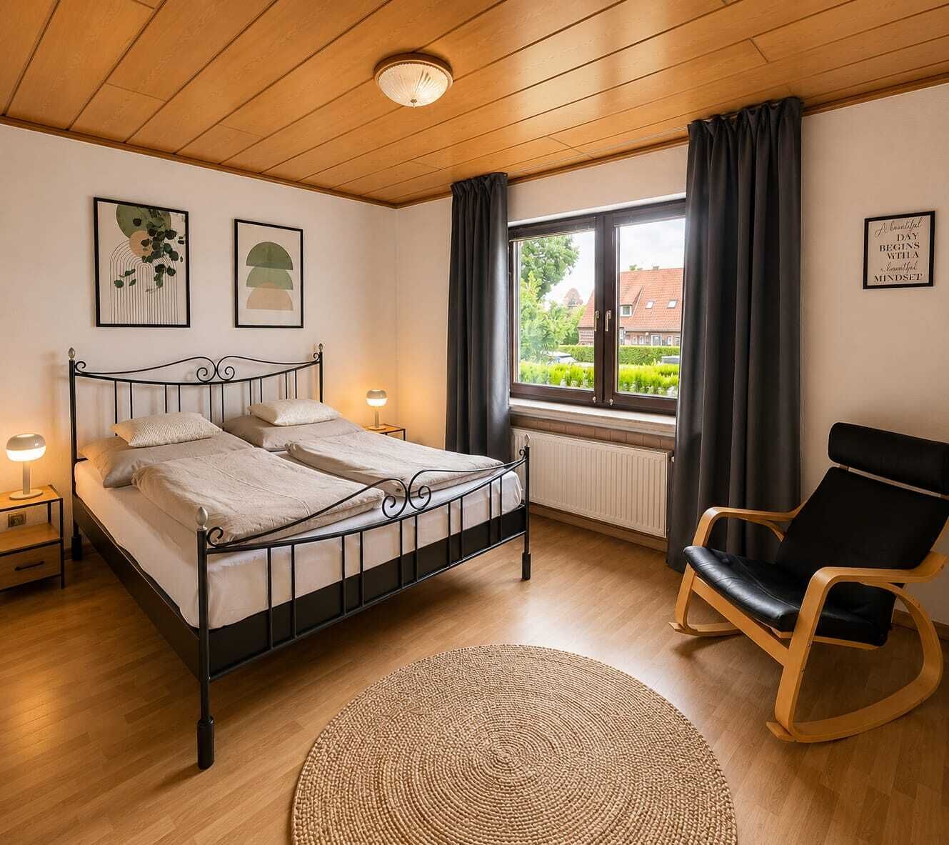 A bedroom with a bed, two bedside lamps, a chair, a window with curtains, and a radiator.