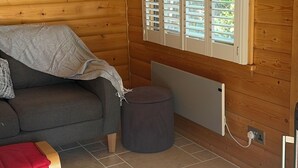 Ein gemütlicher Raum mit Holzvertäfelung, einem grauen Sofa, einem kleinen runden Hocker und einem Fenster mit Rollläden.