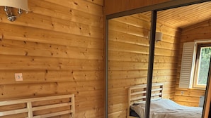 Ein Schlafzimmer mit Holzwänden, einem Bett mit weißer Matratze, einem Nachttisch und einem Spiegel, der ein weiteres Bett reflektiert.