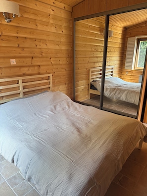 Un dormitorio con paredes de madera, una cama con colchón blanco, una mesita de noche y un espejo que refleja otra cama.