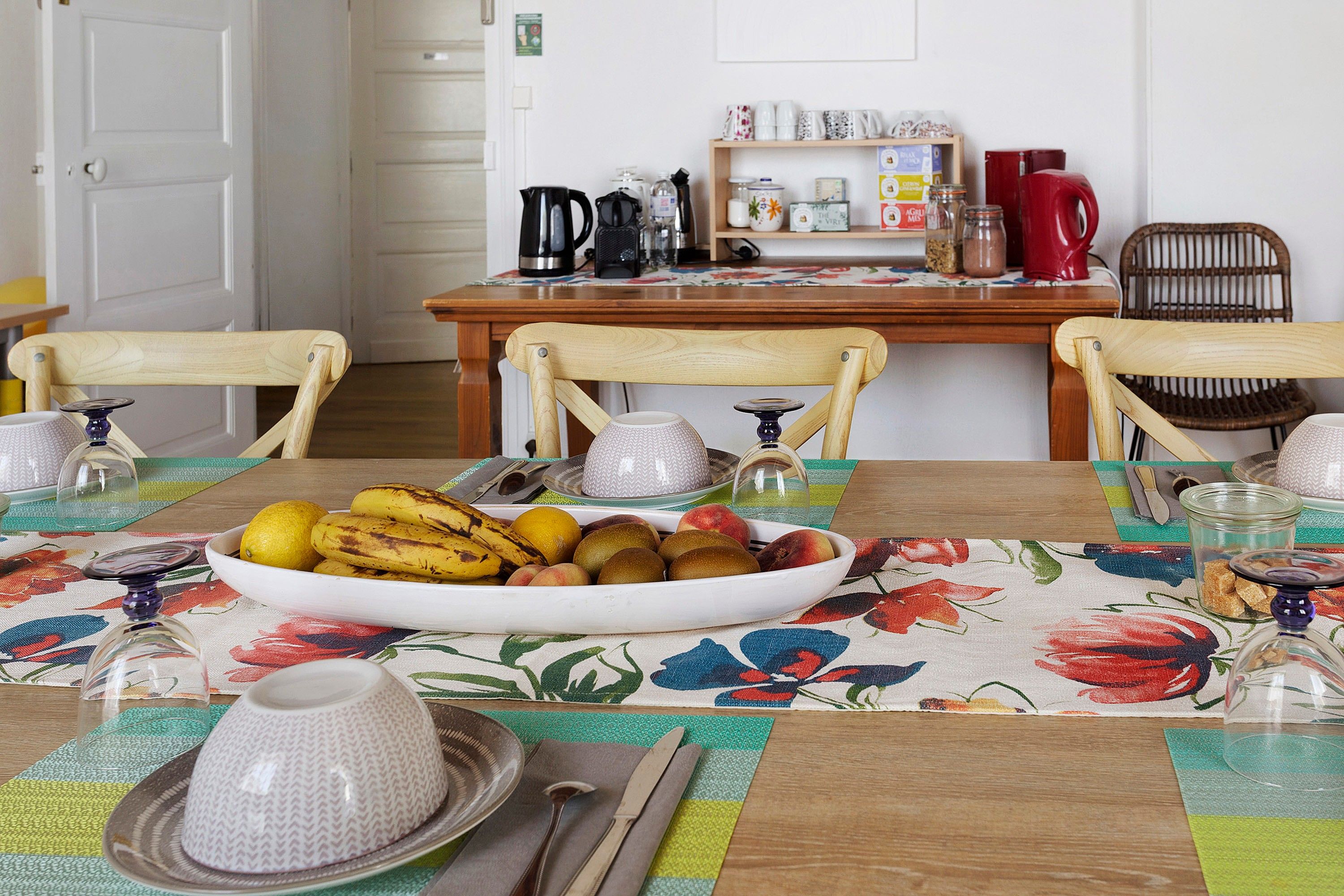 Une table à manger avec un panier de fruits, des couverts et une nappe à motifs floraux.