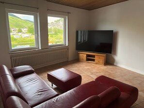Una sala de estar con un televisor de pantalla plana grande, un sofá de cuero rojo y vistas a un paisaje verde a través de dos ventanas.