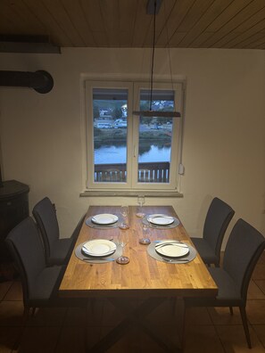 Una mesa de comedor dispuesta para cuatro personas, con sillas, una ventana que ofrece vistas a un cuerpo de agua y casas, y un techo con una lámpara.