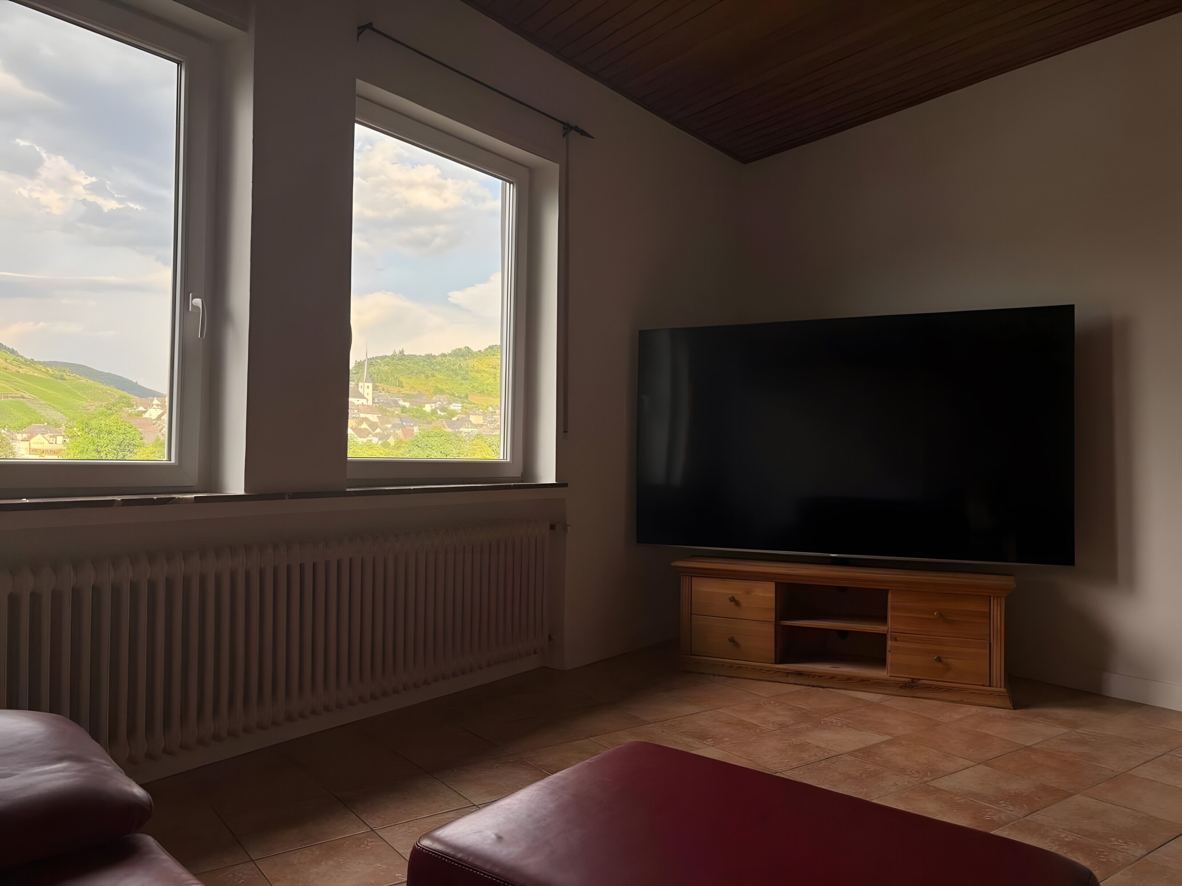 Une pièce dotée d’un grand téléviseur à écran plat posé sur un meuble en bois, de deux fenêtres donnant sur un paysage verdoyant, d’un radiateur placé sous les fenêtres et d’un banc rouge.
