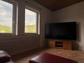 A room with a large flat-screen TV on a wooden stand, two windows with a view of a green landscape, a radiator under the windows, and a red bench.
