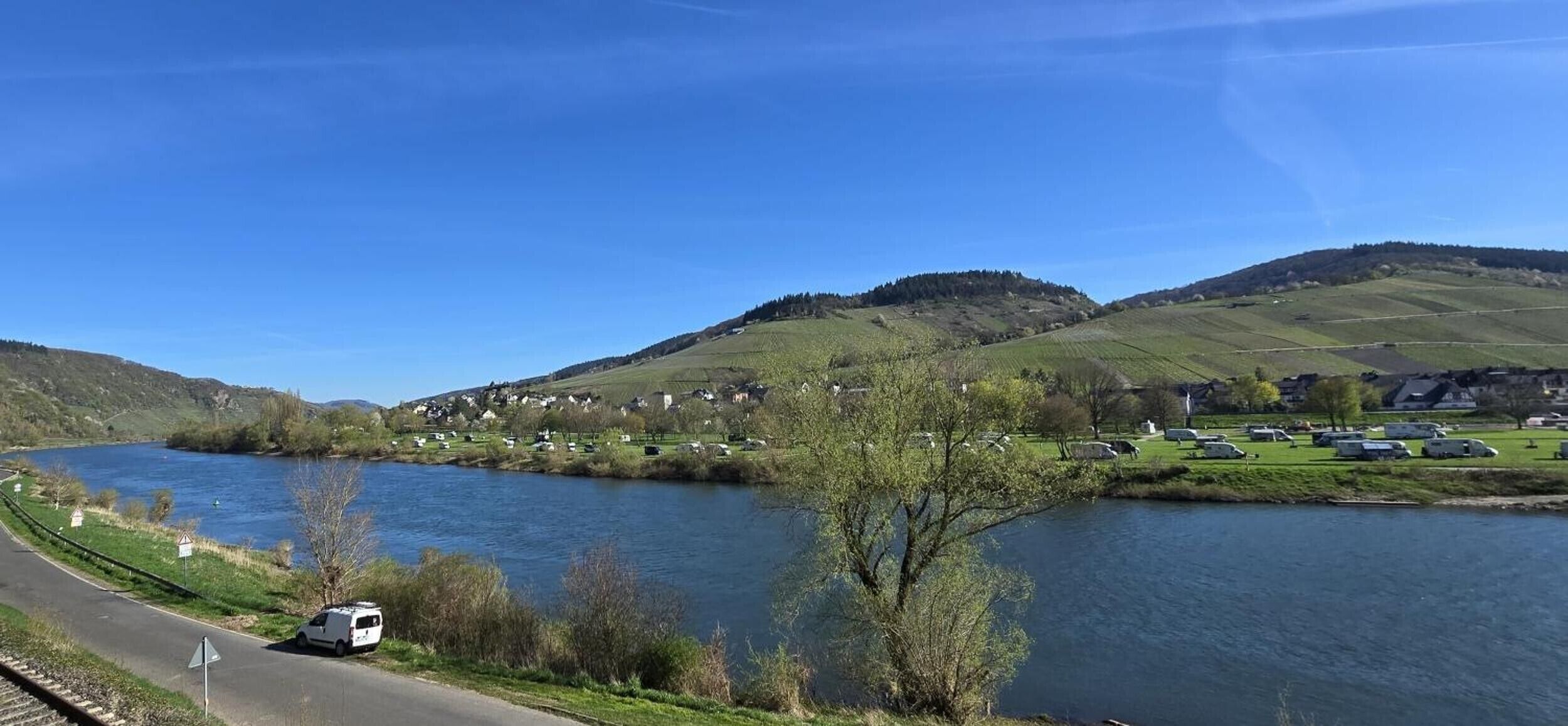 Un río con un pueblo en la orilla opuesta, una carretera con una furgoneta y colinas al fondo.