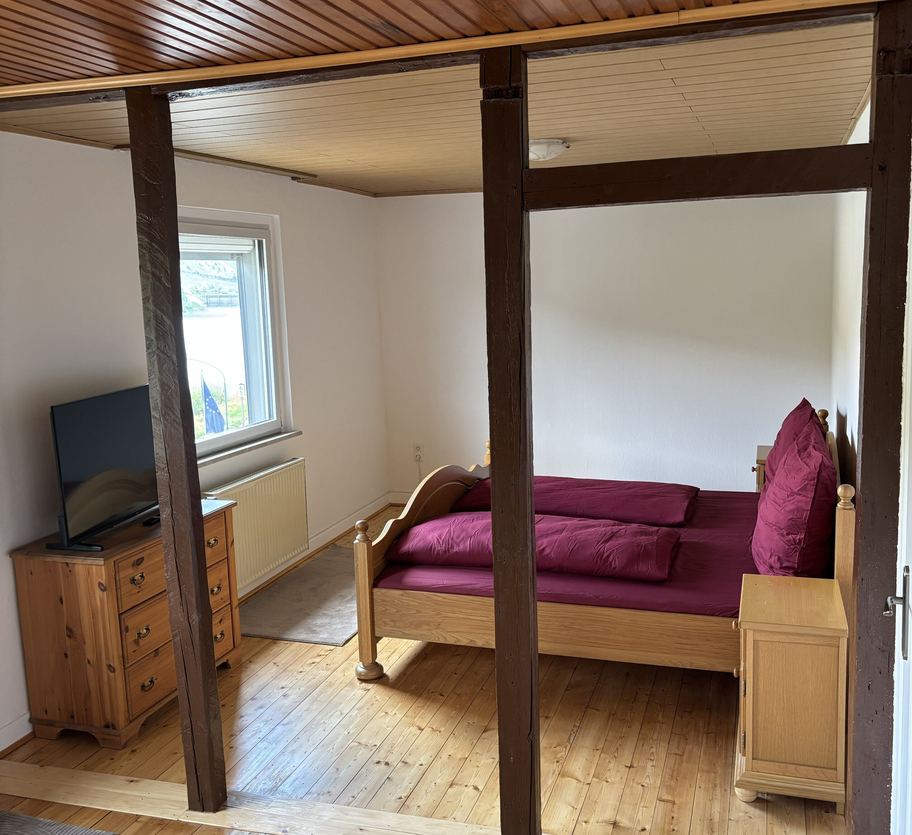 Une chambre à coucher avec un lit à baldaquin en bois, un téléviseur posé sur une commode en bois et une fenêtre donnant sur un paysage verdoyant.