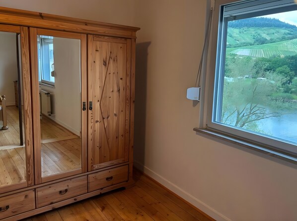 A wooden wardrobe with a mirror, a window with a view of a green landscape, and a thermostat on the wall.