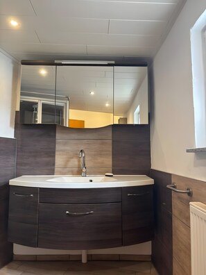 Un baño moderno con un espejo grande, muebles de madera oscura, un lavamanos blanco y una zona de ducha visible al fondo.