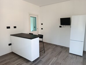 Una cuina compacta amb taulell blanc, un frigorífic i un televisor penjat a la paret.