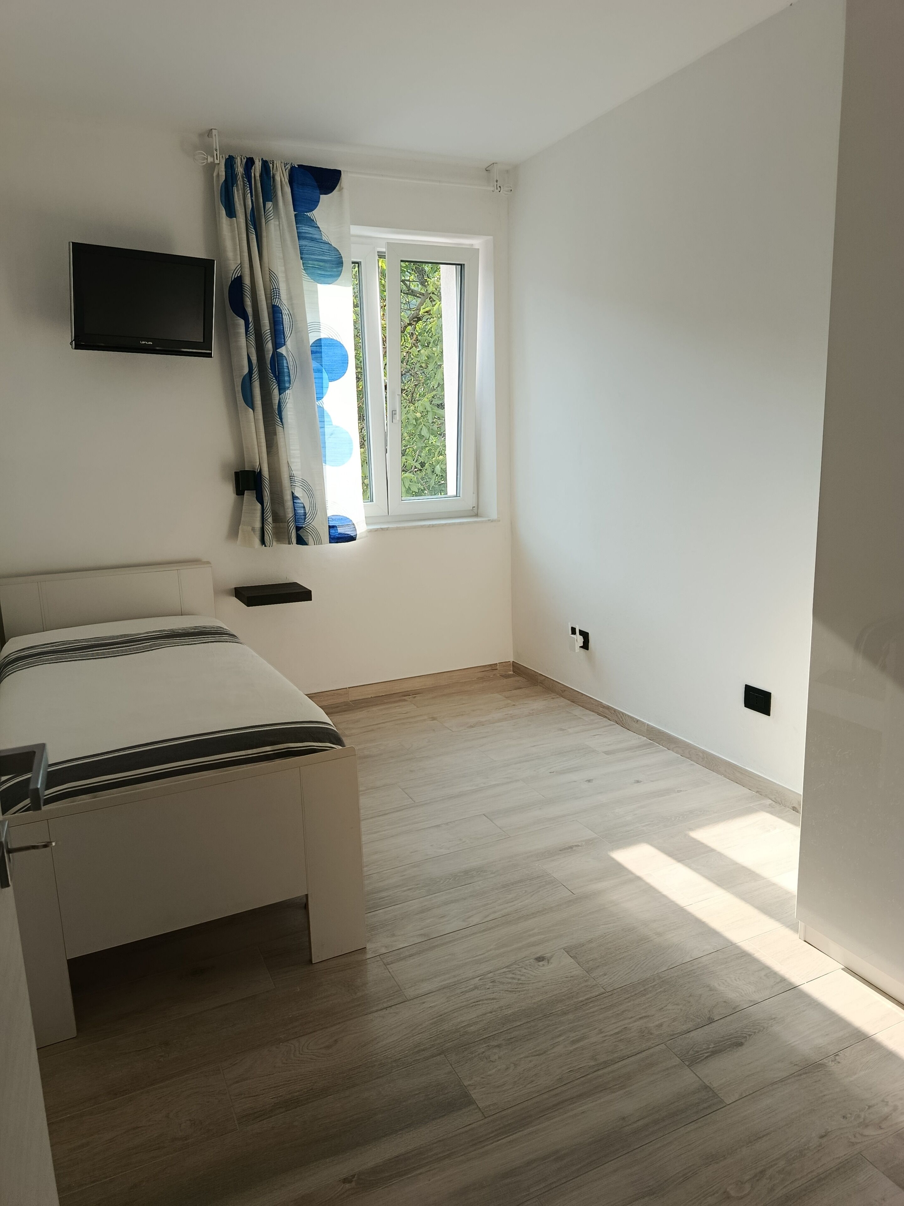 A bedroom with a bed, a TV mounted on the wall, a window with curtains, and a door.