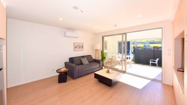 A modern living room with a grey sofa, a coffee table, and a small side table. There is a sliding glass door leading to an outdoor area with a table and chairs. - The Cosy Nook for a Relaxing City Stay (Christchurch)