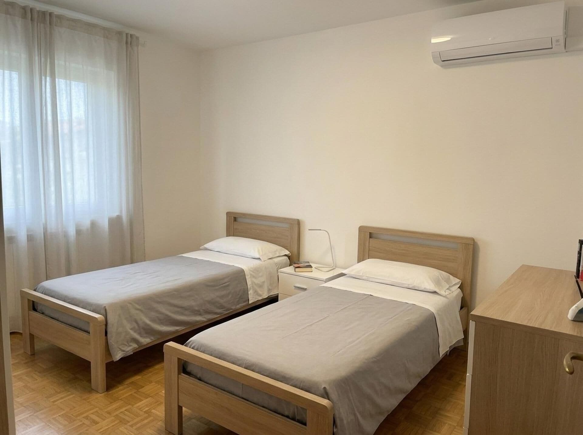 A room with two single beds, a bedside table with a lamp, a small desk, and an air conditioning unit.