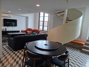 A modern living room with a black sofa, a round table with chairs, a flat-screen TV, and a spiral staircase. - Junto a la histórica Muralla del Baluarte | 1BR + 2BA | Hasta 6 huéspedes (Panamá)