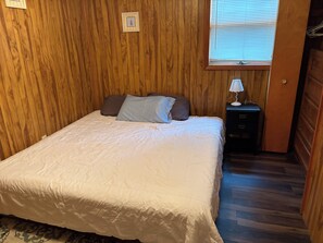 Un dormitorio con pared de paneles de madera, cama con ropa blanca, mesita de noche con lámpara y ventana con persianas.