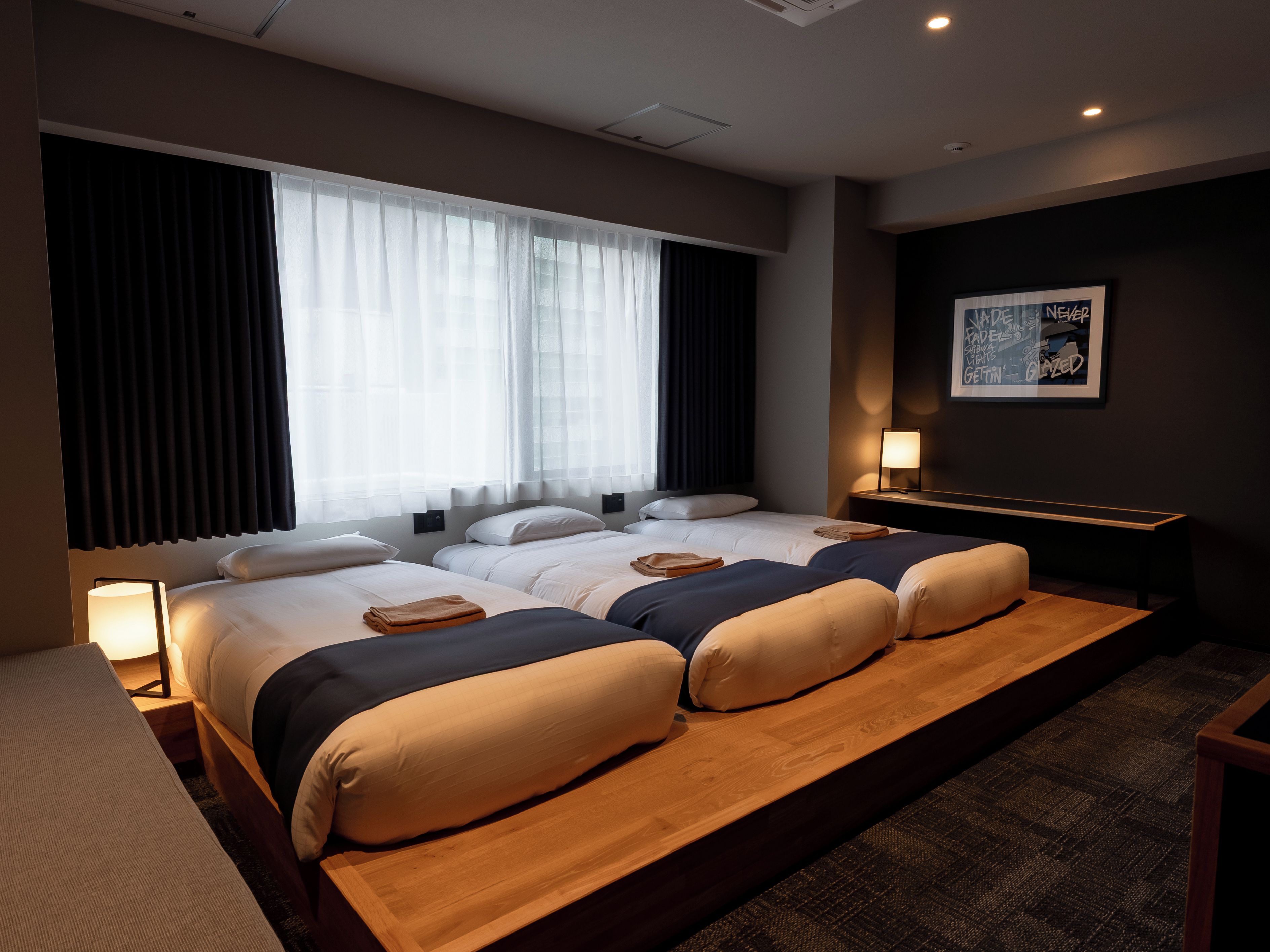 A hotel room with two beds, a large window with sheer curtains, a wall-mounted artwork, and a bedside table with a lamp.