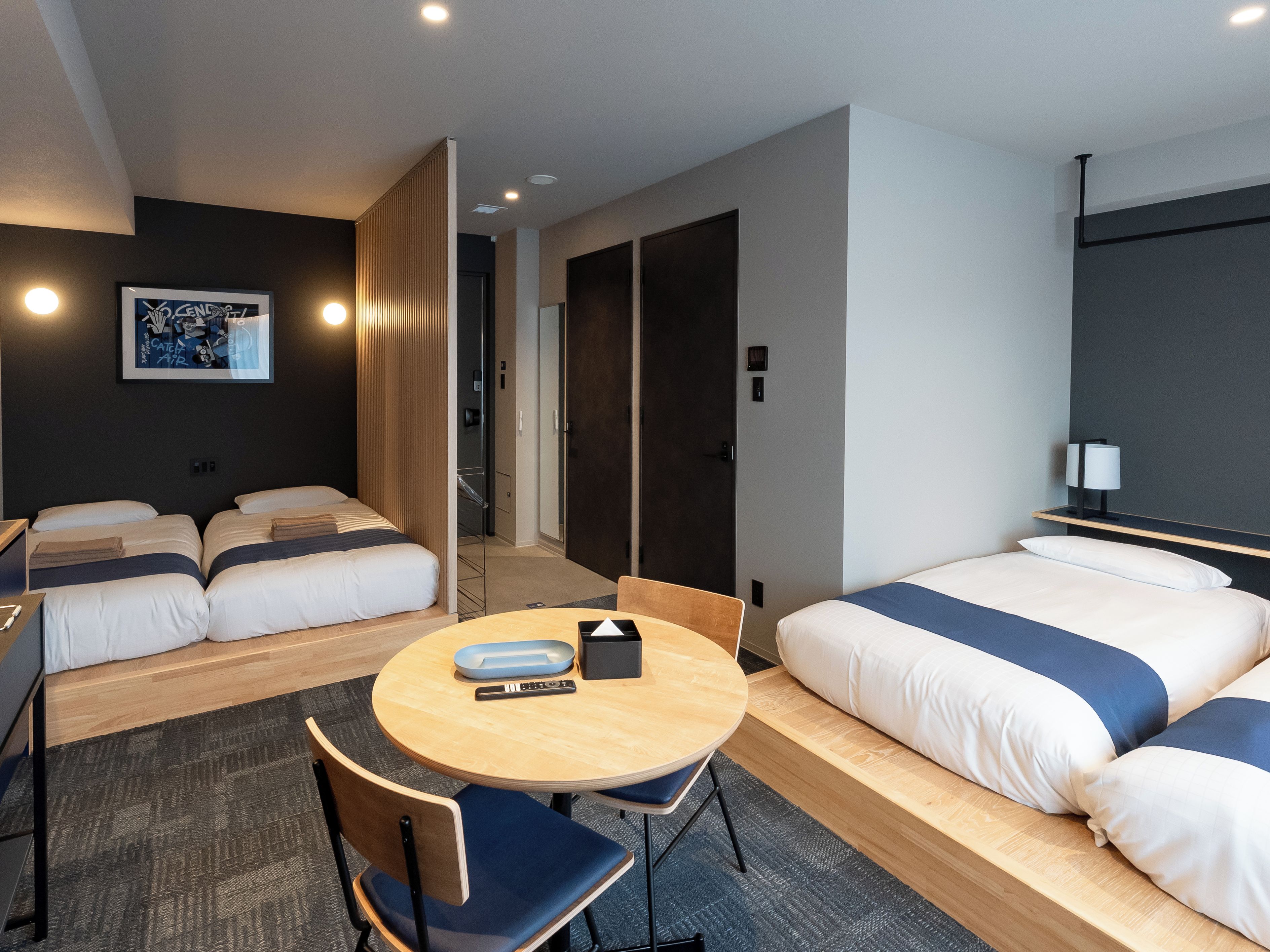 A hotel room with two beds, a round table with chairs, a small desk, and a wall-mounted lamp.