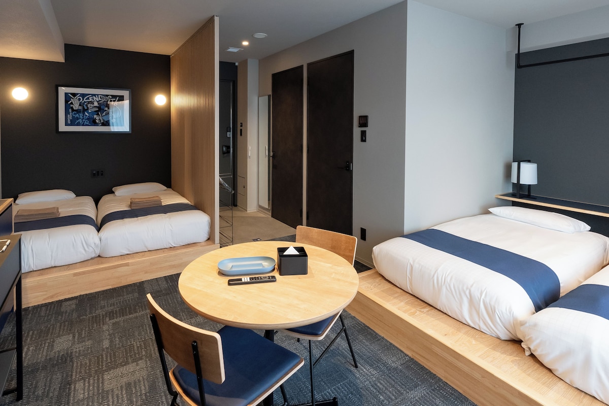 Habitación de hotel con dos camas, una mesa redonda con sillas, un escritorio pequeño y una lámpara de pared.