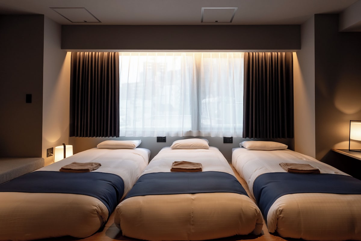 Una habitación de hotel con tres camas, un ventanal con cortinas y suelo de madera.