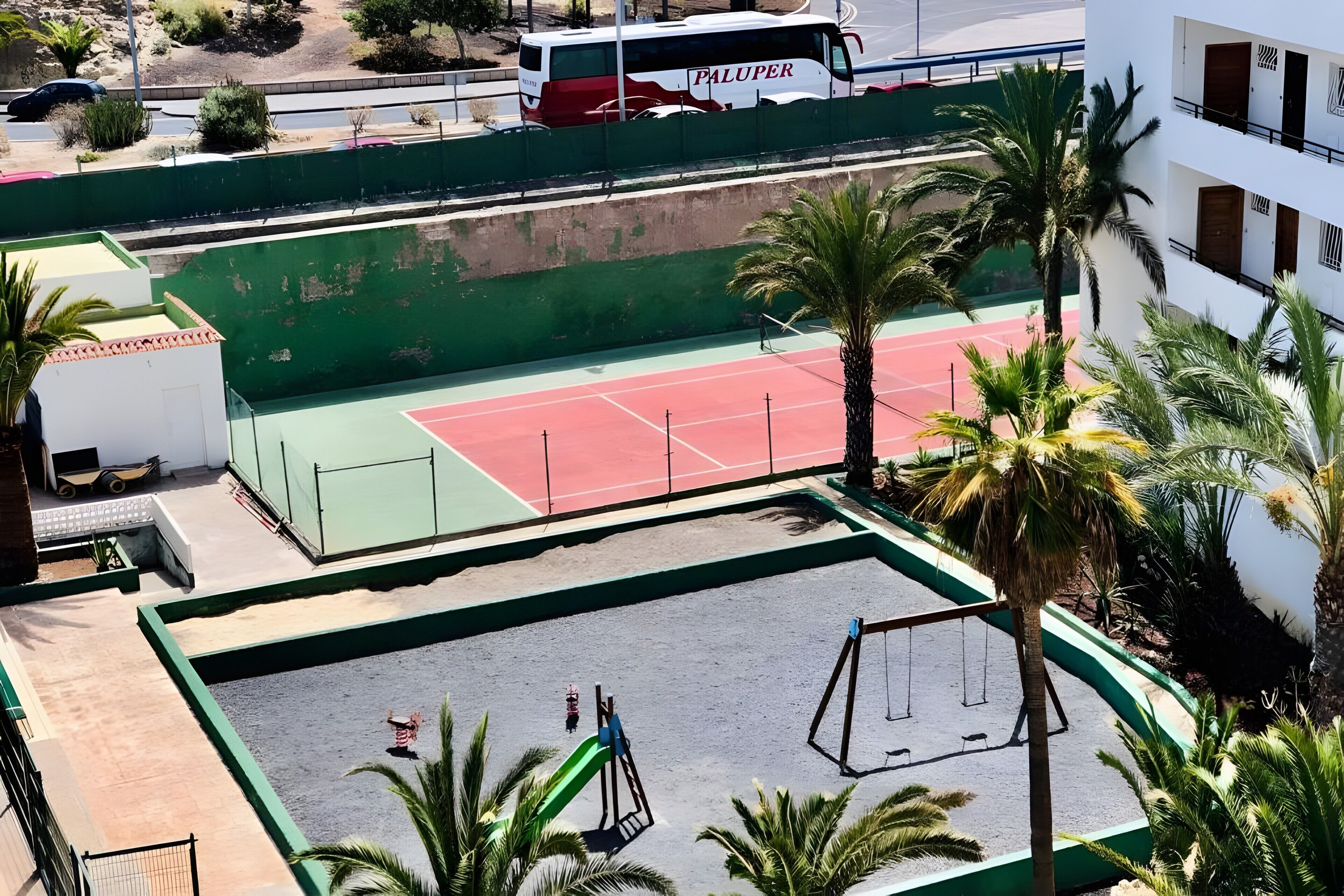 Een tennisbaan met een net, omringd door palmbomen en een groene hekken.