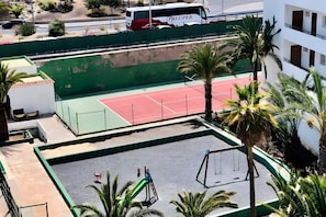 Een tennisbaan met een net, omringd door palmbomen en een groene hekken.