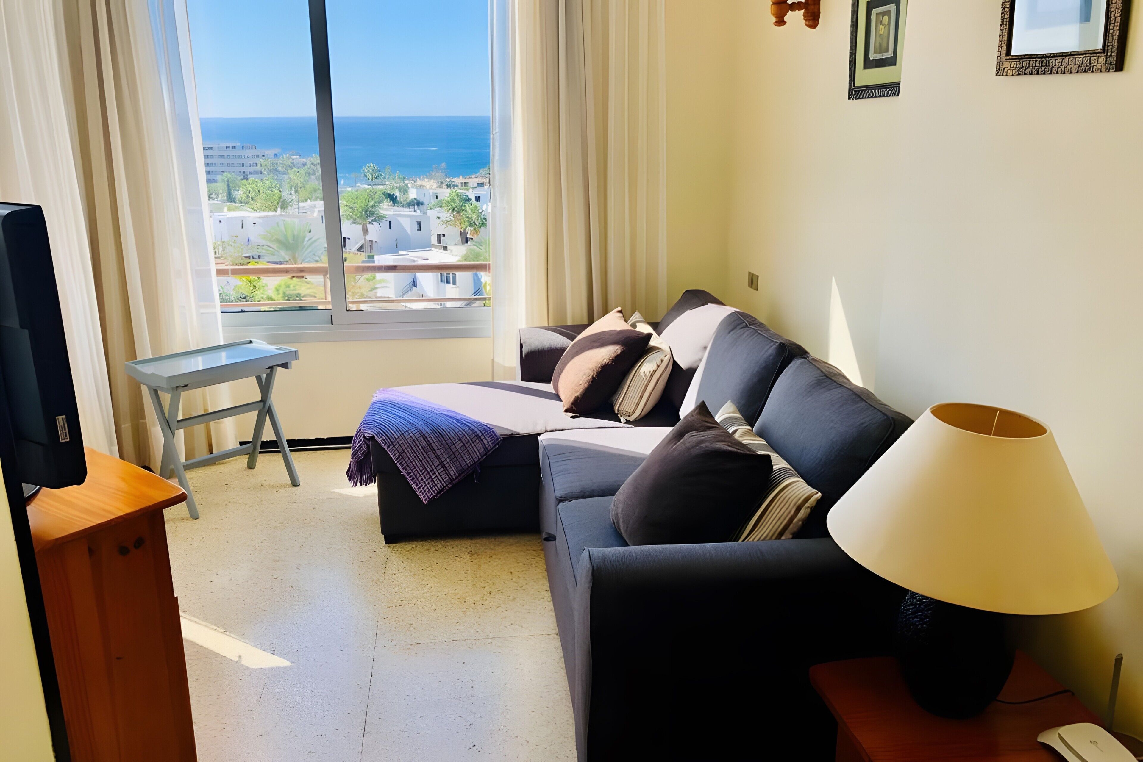 A living room with a sofa, a small table, a lamp, and a view of the sea through a large window.