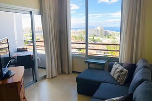 A living room with a sofa, a table, and a view of a cityscape through large windows. - Vacation Apartment 'Cozy  Playa Las Americas' with Sea View, Balcony and Wi-Fi (Costa Adeje)
