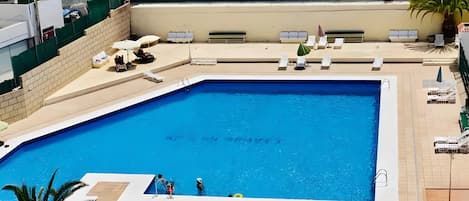 A rectangular swimming pool with a tiled deck surrounding it, equipped with lounge chairs and umbrellas.