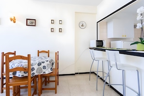 Een compacte keuken met een eethoek, met een houten tafel met een tafelkleed met een patroon, een witte koelkast, een magnetron en een klok aan de muur.
